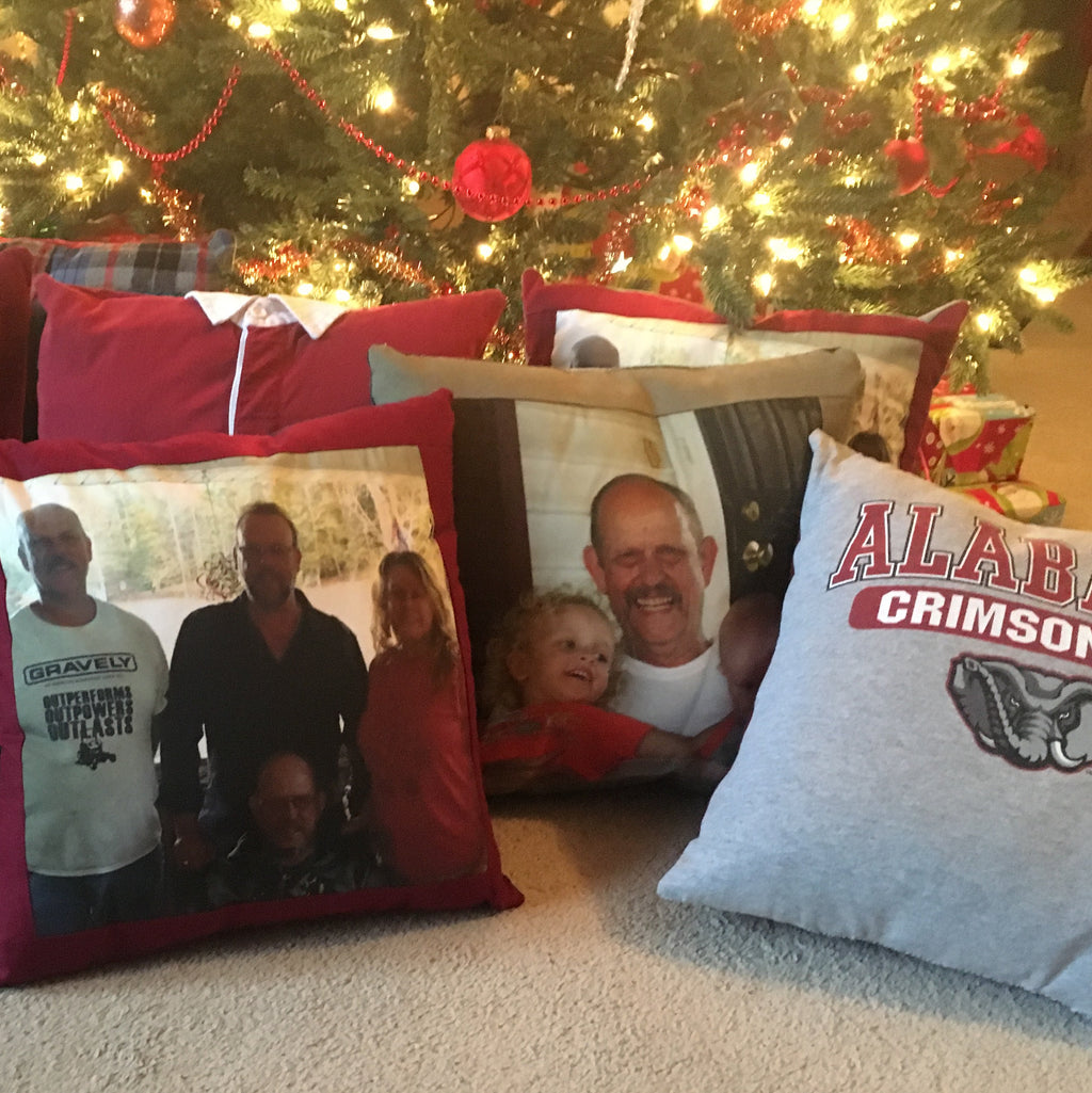 A group of custom photo pillows arranged on a beige carpet in front of a decorated Christmas tree. The pillows feature various fabric patterns including solid red, plaid, and heather gray, with printed family photos and one pillow showing the Alabama Crimson Tide logo.