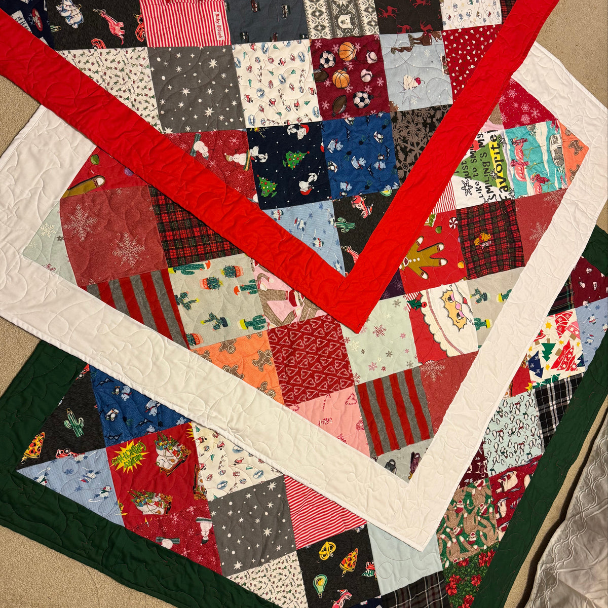 A handmade Christmas-themed patchwork quilt laid out on a beige carpeted floor. The quilt features a vibrant red V-shaped border, a white border, a dark green border, and a central patchwork grid of various colorful fabric squares with holiday motifs like Santa Claus, snowflakes, and pine trees. This quilt is made out of children's pajamas.