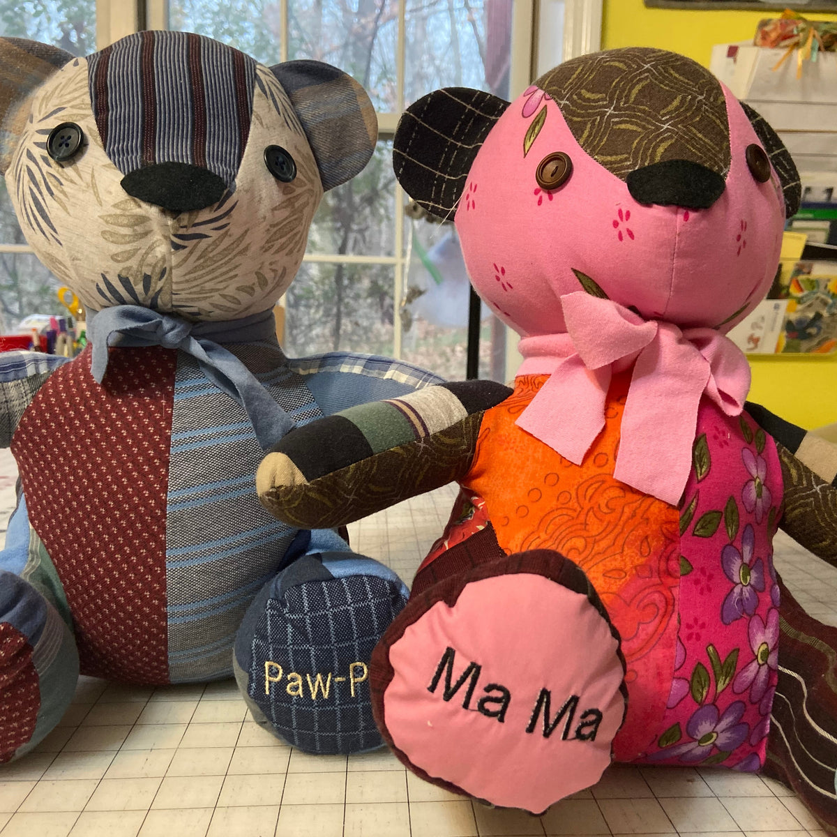 Two handmade patchwork teddy bears sitting side by side on a table. The left bear has blue, gray, and red patchwork fabric with a blue bow.