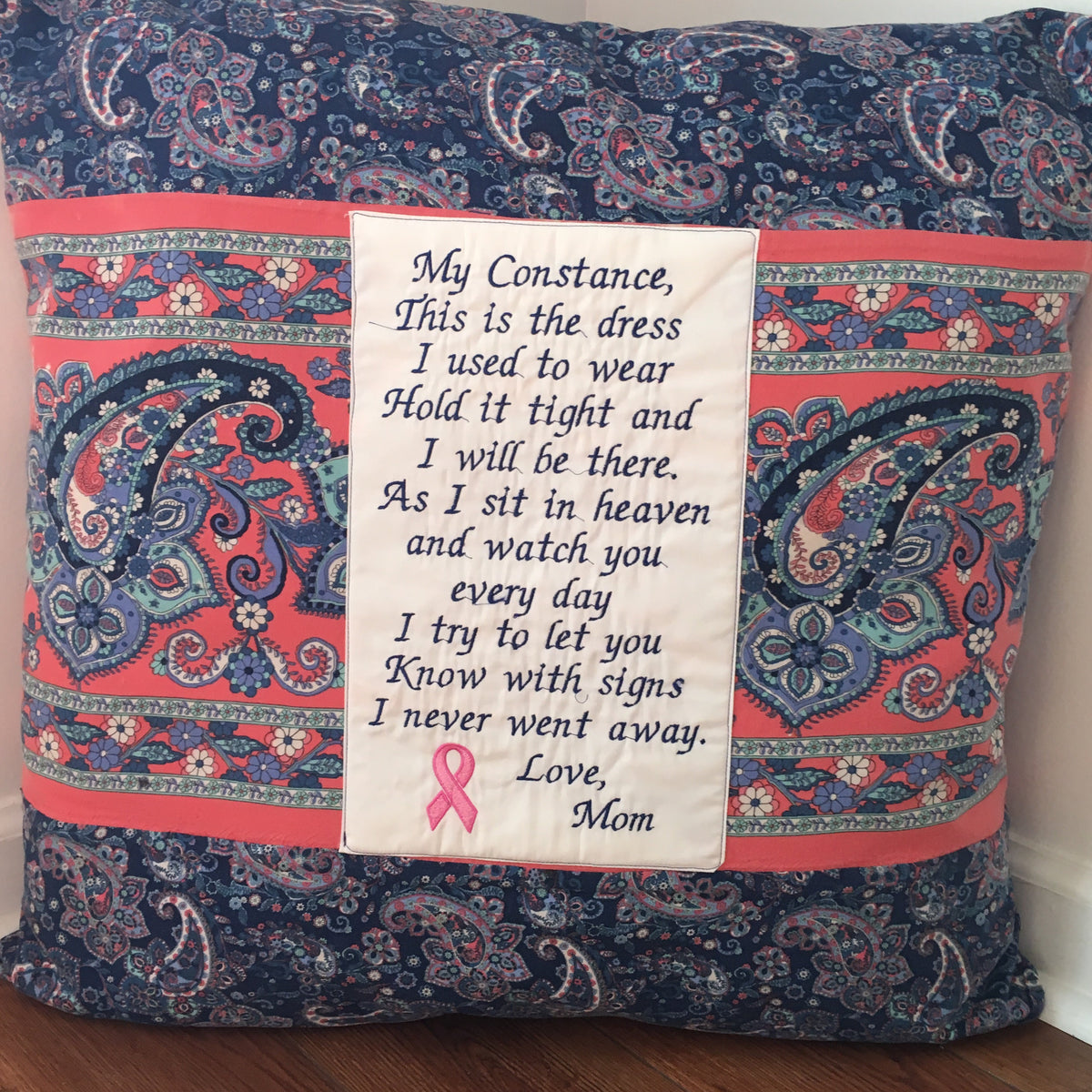 A square decorative throw pillow with a navy blue paisley pattern, coral pink borders, and a central white fabric panel printed with a memorial poem addressed to 'Mom' with a pink ribbon symbol.