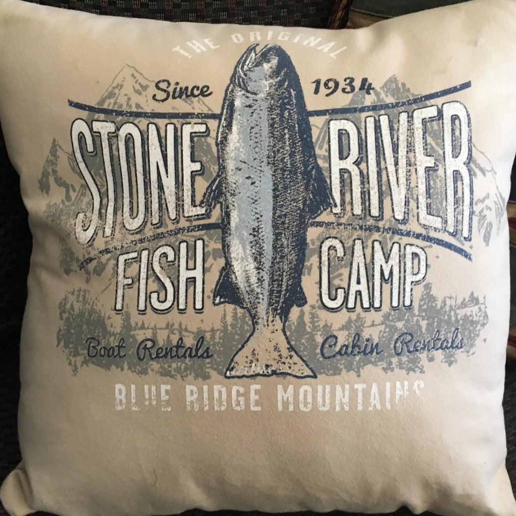 A square throw pillow with a vintage-style 'Stone River Fish Camp' graphic featuring a fish illustration, resting on a dark woven couch.