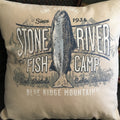 A square throw pillow with a vintage-style 'Stone River Fish Camp' graphic featuring a fish illustration, resting on a dark woven couch.