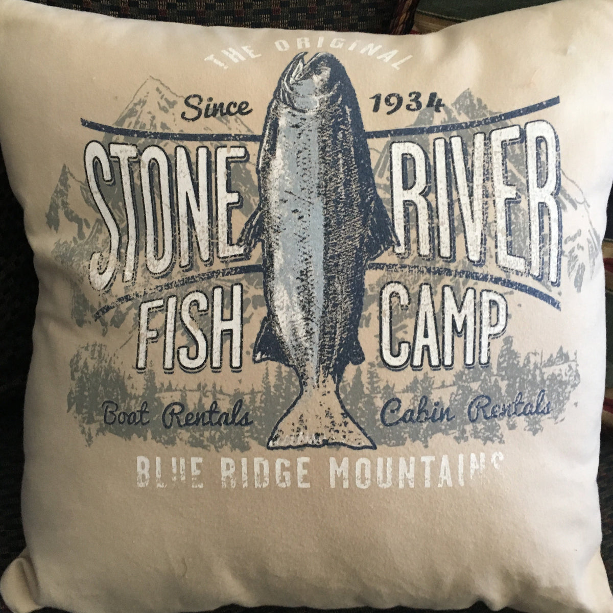 A square throw pillow with a vintage-style 'Stone River Fish Camp' graphic featuring a fish illustration, resting on a dark woven couch.