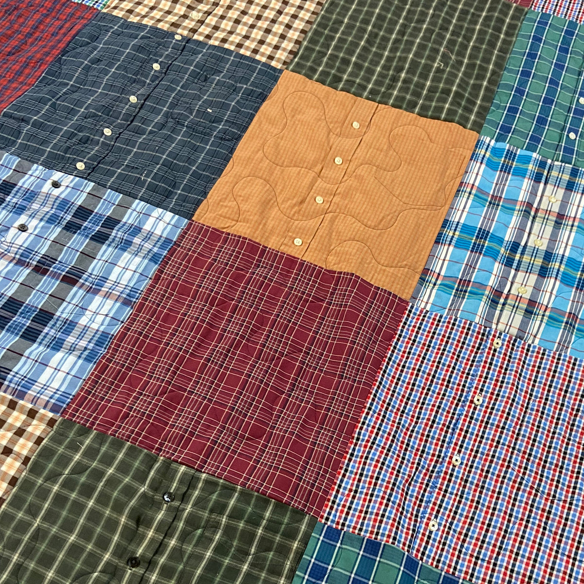 A handmade quilted patchwork blanket made from vintage plaid button-down shirts, laid out on a beige carpet. The quilt features various colors and patterns including blue, green, red, and brown plaid, with a personalized patch reading 'Ernest Campbell Jr.