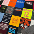A colorful patchwork quilt made from various high school and college athletic t-shirts, featuring vibrant panels of red, yellow, orange, blue, black, and white with printed sports logos and slogans. A gray quilted fabric item is partially visible in the bottom right corner.