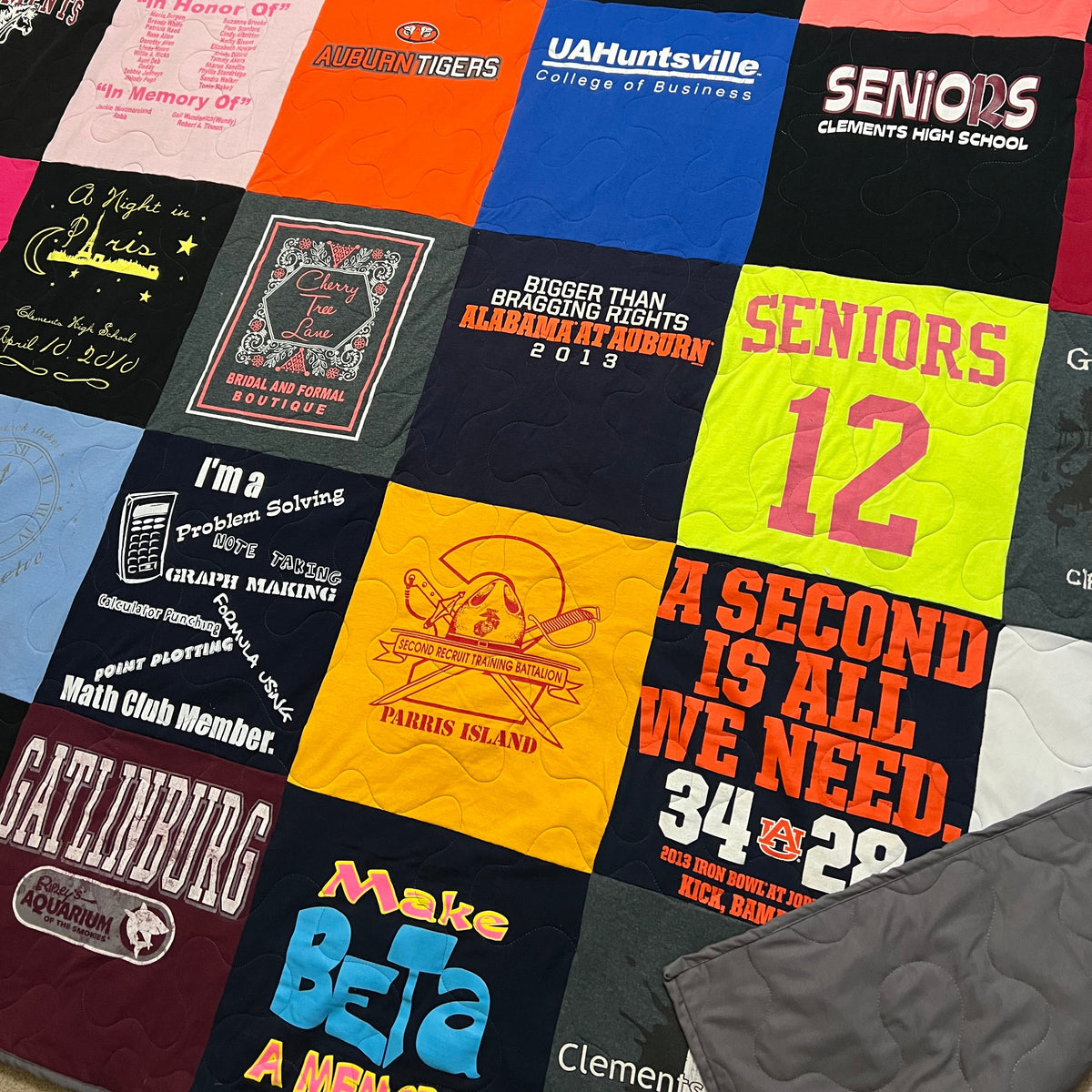 A colorful patchwork quilt made from various high school and college athletic t-shirts, featuring vibrant panels of red, yellow, orange, blue, black, and white with printed sports logos and slogans. A gray quilted fabric item is partially visible in the bottom right corner.
