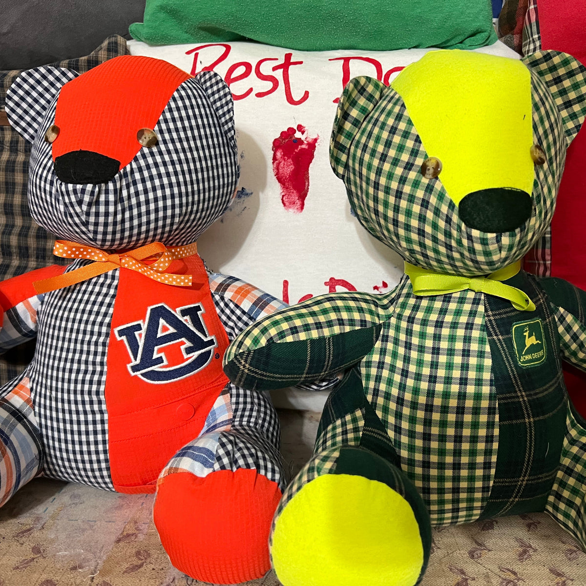 Two handmade patchwork teddy bears made from plaid and solid fabrics, posed together on a cushioned surface with pillows behind them. One bear features an Auburn Tigers 'AU' logo, and the other features a green John Deere logo patch.