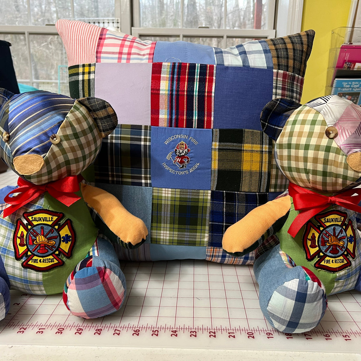 A handmade patchwork fabric pillow with a central fire department emblem, flanked by two matching patchwork fabric teddy bears wearing fire department patches and red bows, set on a measuring mat in front of a window.