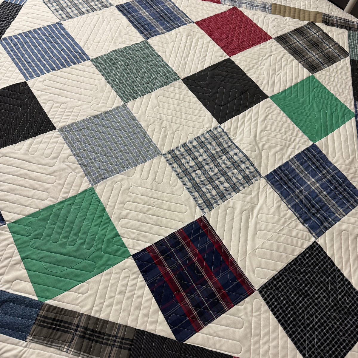 A handmade patchwork quilt laid flat on a table, featuring a grid pattern of square panels in various colors including blue, green, red, black, and gray, with some plaid and striped designs. The quilt is detailed with quilting stitches.