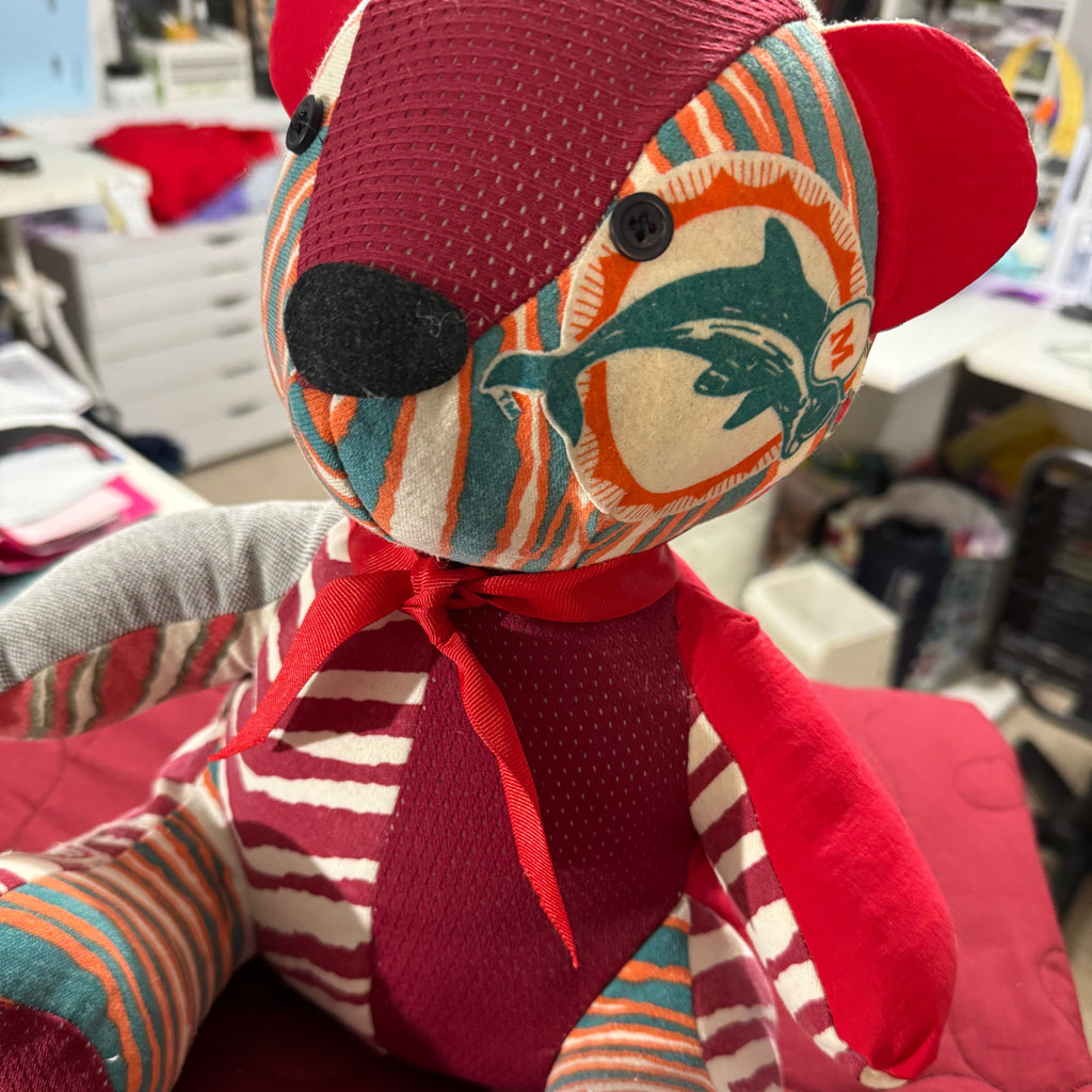 A handmade patchwork teddy bear plush toy made from various colorful fabrics, including red mesh, striped teal and cream, and orange and red patterns, sitting on a red quilted blanket in a cluttered room.
