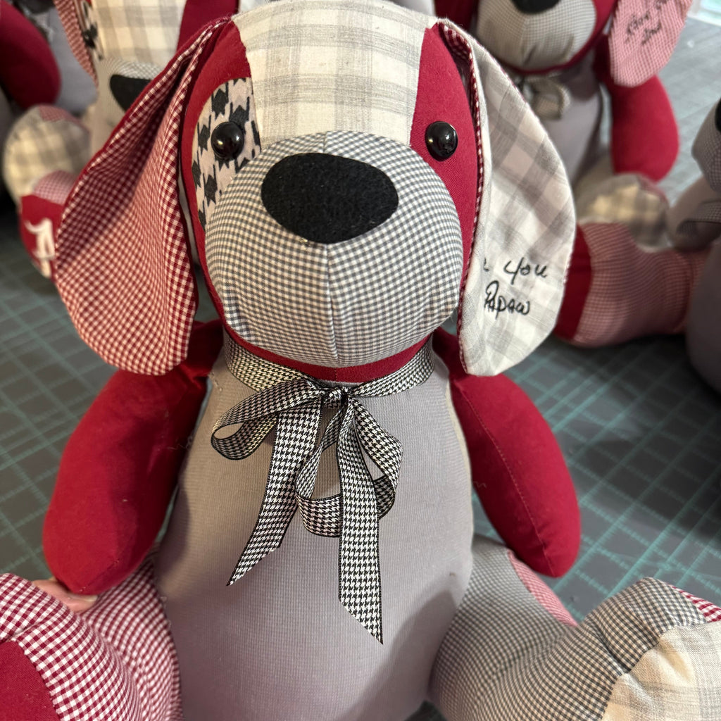 A handmade patchwork fabric dog plush toy with red and gray fabric panels, a black-and-white gingham patterned face and ears, a red collar with a black-and-white bow, and embroidered text on one ear, sitting on a gray cutting mat.