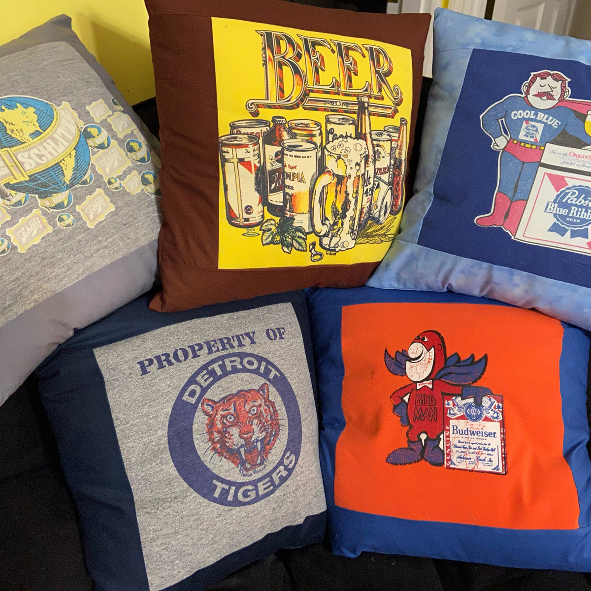 Five decorative throw pillows arranged on a dark couch, each featuring a different vintage-style graphic print including beer cans, a globe, a Pepsi character, a Detroit Tigers tiger head, and a red bird mascot.