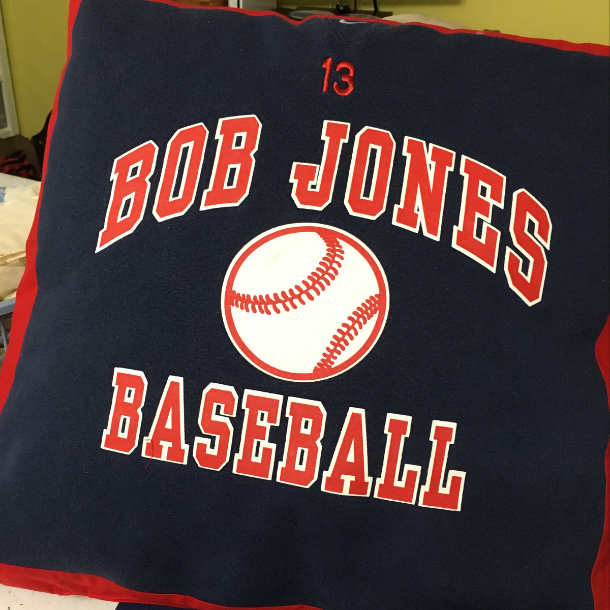 A custom navy blue throw pillow with red trim, featuring the text 'BOB JONES BASEBALL' and the number '13' in red varsity lettering around a baseball graphic.