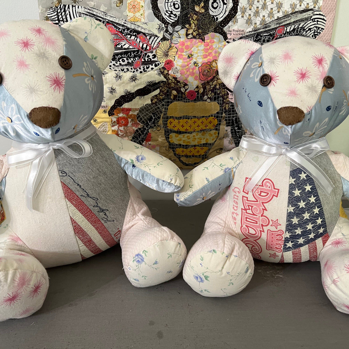 Two handmade patchwork fabric teddy bears sitting side by side on a gray surface, with a colorful quilted owl decoration in the background.