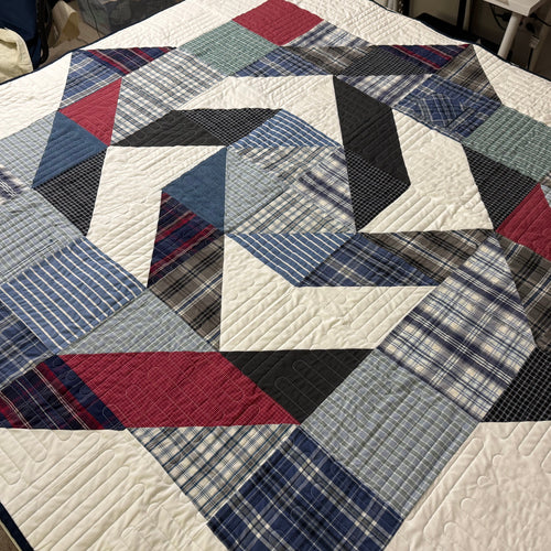 A handmade geometric quilt with a star-like pattern, made from various blue, red, gray, and black plaid and striped fabrics, laid out on a table in a sewing room.