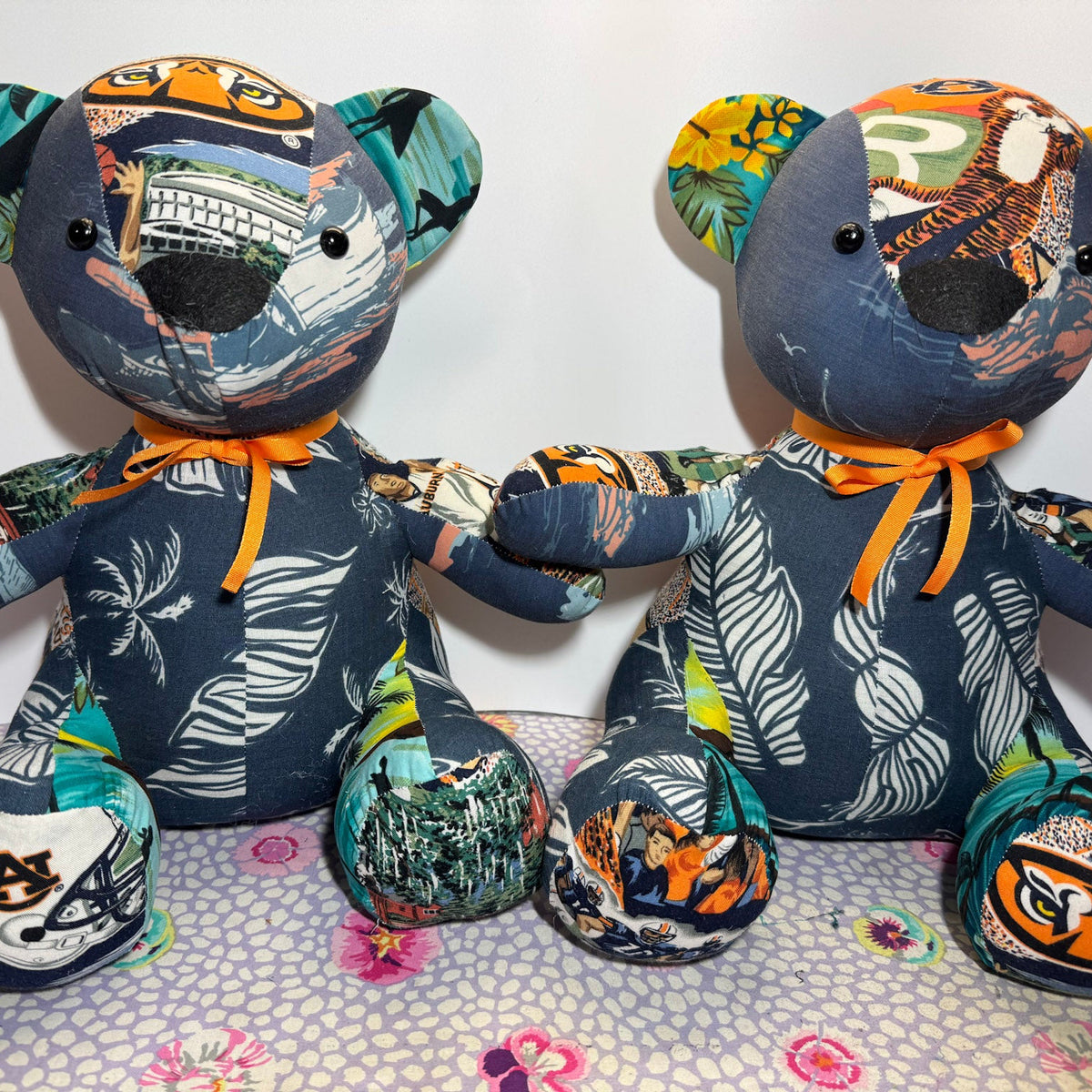 Two handmade patchwork fabric teddy bears sitting side by side, holding hands. The bears are made from various colorful patterned fabrics including tropical prints, cityscapes, and abstract designs, with dark blue/navy fabric on their main bodies and bright orange ribbon neckties.